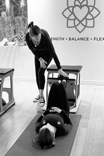 An instructor guiding a client lying on a mat with her feet on a Pilates chair pedal