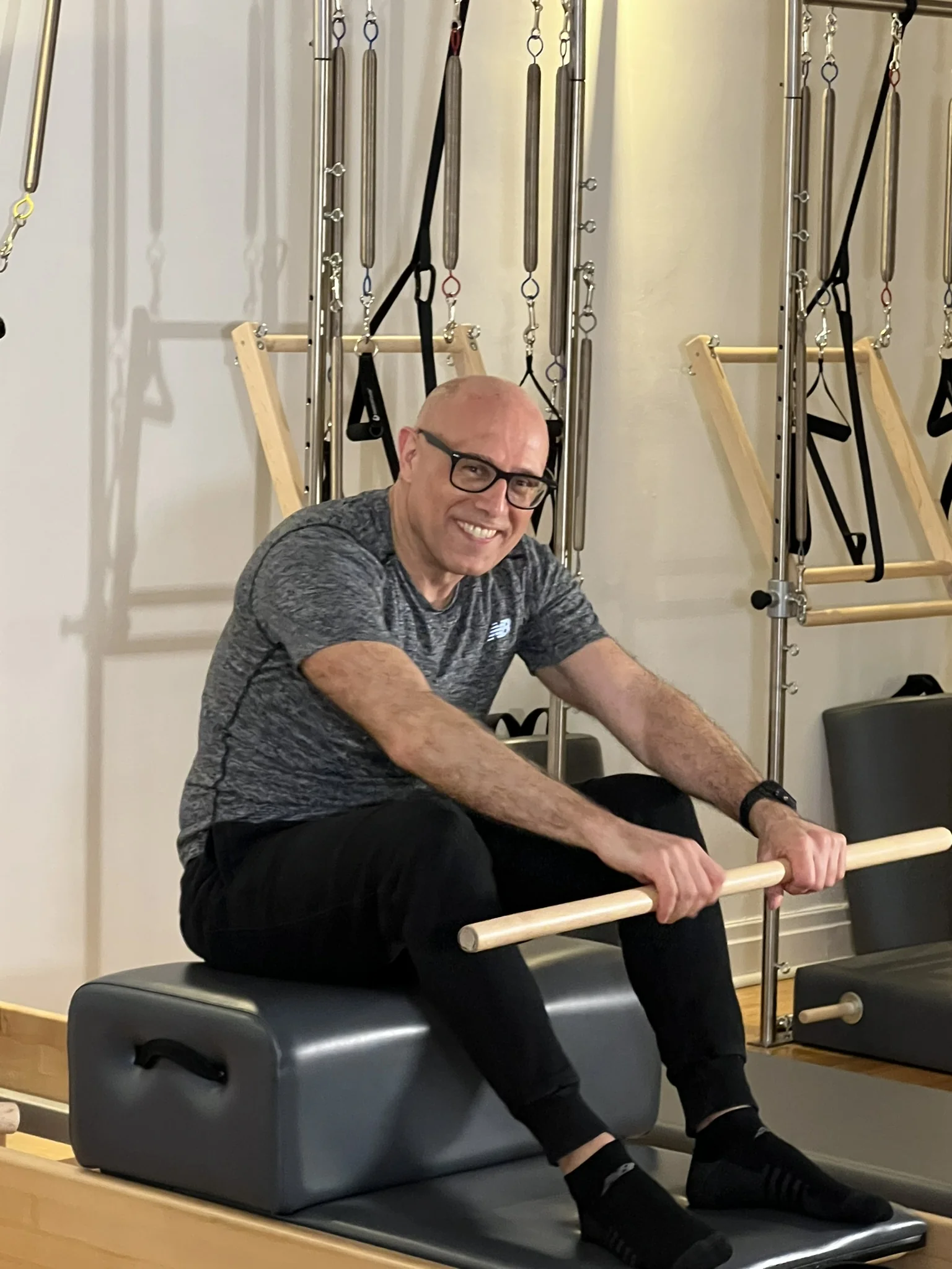 A smiling man seated on a Reformer box holding a wooden bar