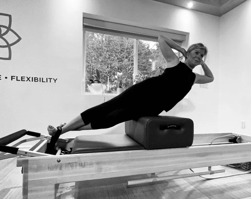 A woman practising a side bend over a Reformer box with her feet secured by a strap